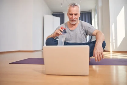 Older man sitting on a yoga mat holding a water bottle, looking at a laptop screen in a bright room.